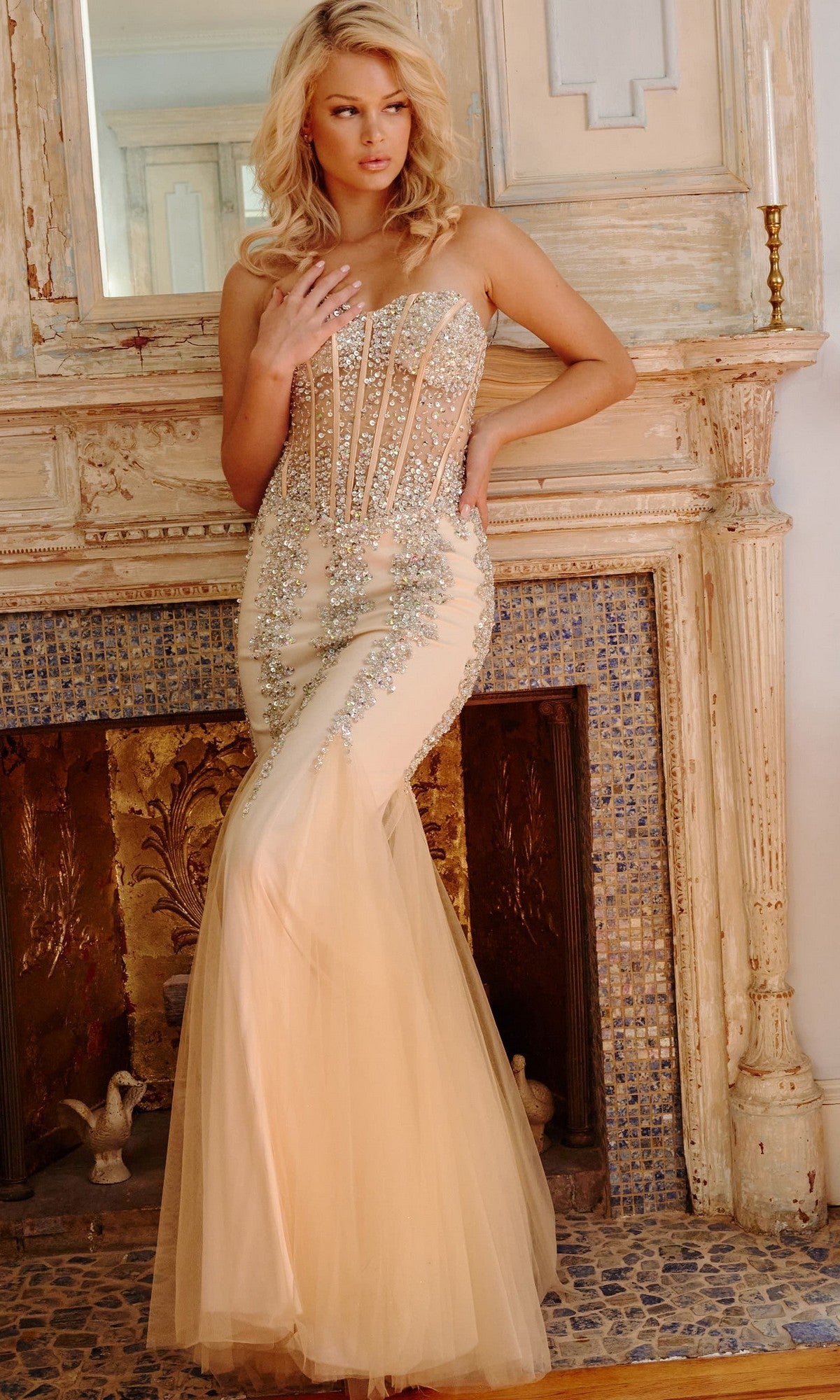 A woman wearing the Long Strapless Sweetheart Jovani Prom Dress 5908, featuring sparkling embellishments and a fitted, floor-length silhouette, stands in front of a decorated fireplace.